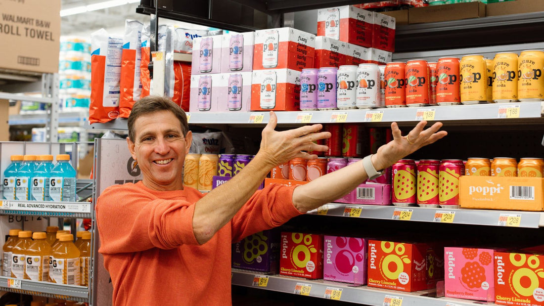 a man smiling in front of a grocery store soda aisle, pointing to the row of colorful culture pop cans