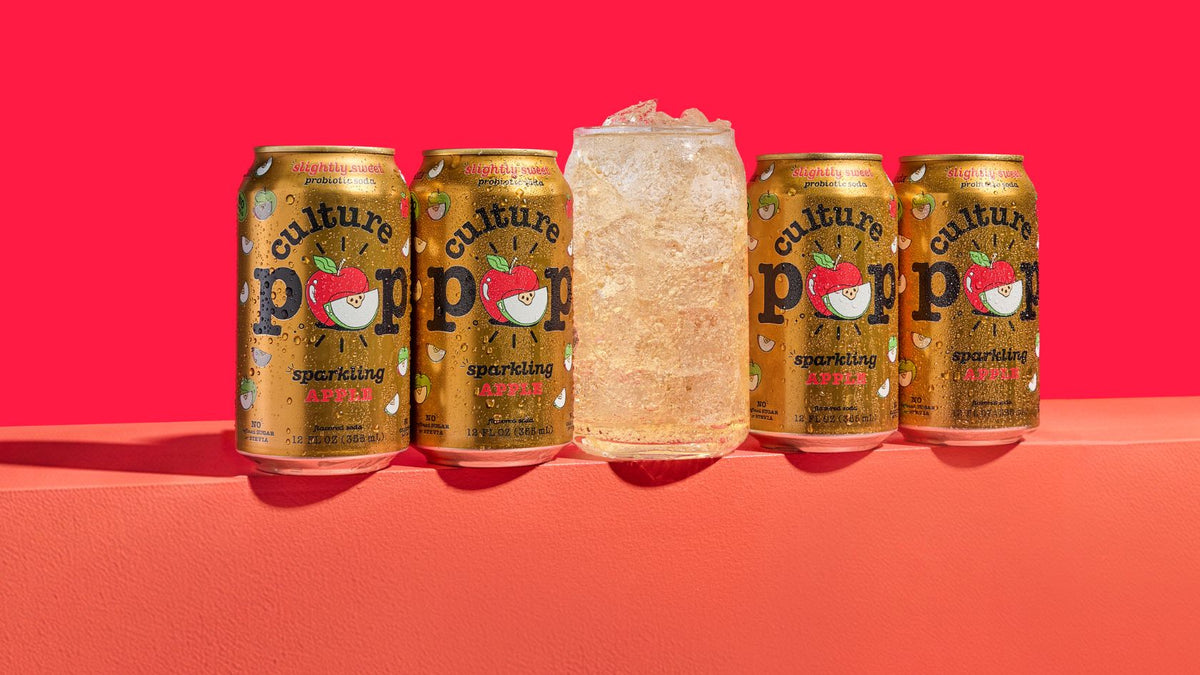 4 gold cans of culture pop soda and a cold glass of apple soda with ice, on a red background