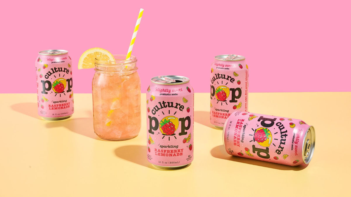 4 cans of raspberry lemonade culture pop pink cans with fruit on the packaging and a mason jar glass with liquid in it on a pink and yellow background